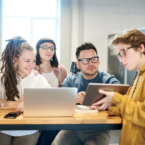 four people are sitting at a table while one of them shows the others something on their laptop